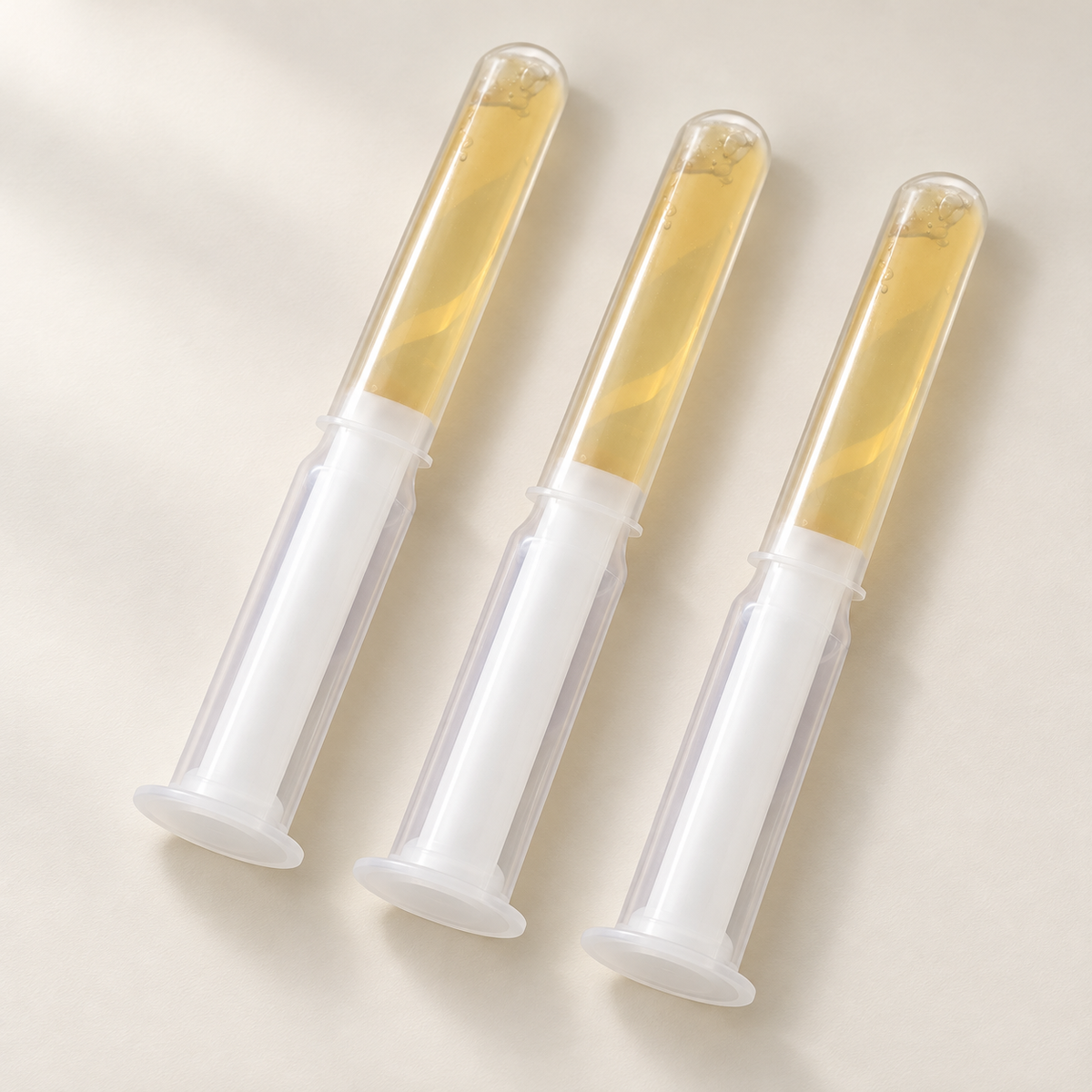 Trois applicateurs Virgin Secret de 10 ml, corps transparent, gel ambré et poussoir blanc, sur fond ivoire.
